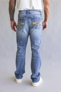 Rear view of a person wearing light blue denim jeans with decorative embroidered patch detailing on the back pockets, paired with a white t-shirt against a neutral gray background.