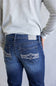 Rear view of a person wearing blue denim jeans with decorative contrast stitching on the back pockets, paired with a light gray long-sleeve top against a plain light gray background.