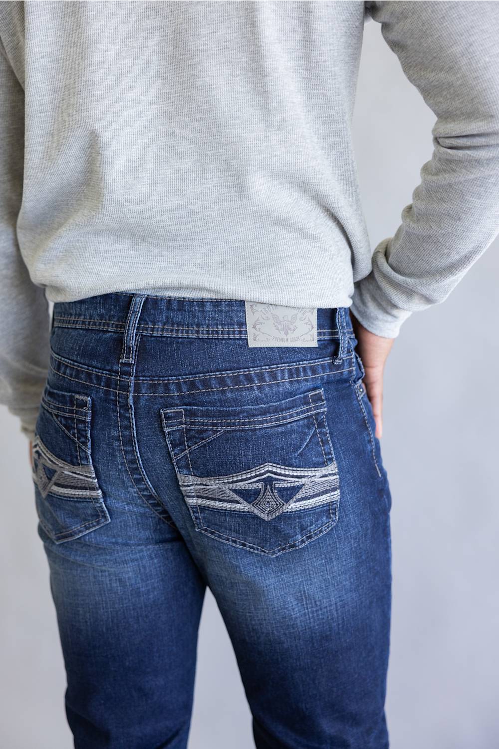 Rear view of a person wearing blue denim jeans with decorative contrast stitching on the back pockets, paired with a light gray long-sleeve top against a plain light gray background.