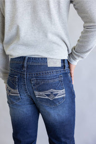 Rear view of a person wearing blue denim jeans with decorative contrast stitching on the back pockets, paired with a light gray long-sleeve top against a plain light gray background.