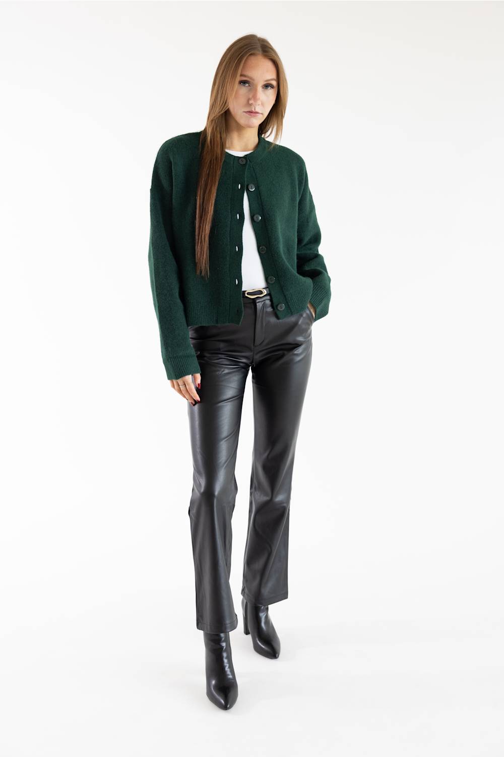 A woman modeling a dark green knit cardigan over a white top, paired with black leather pants and black ankle boots, against a plain white studio background.