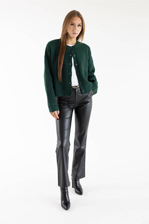A woman modeling a dark green knit cardigan over a white top, paired with black leather pants and black ankle boots, against a plain white studio background.