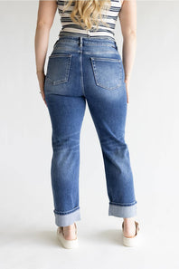 A woman viewed from the back wearing blue denim jeans with a straight-to-medium leg and rolled cuffs, paired with a striped top and white platform sandals, against a plain white studio background.