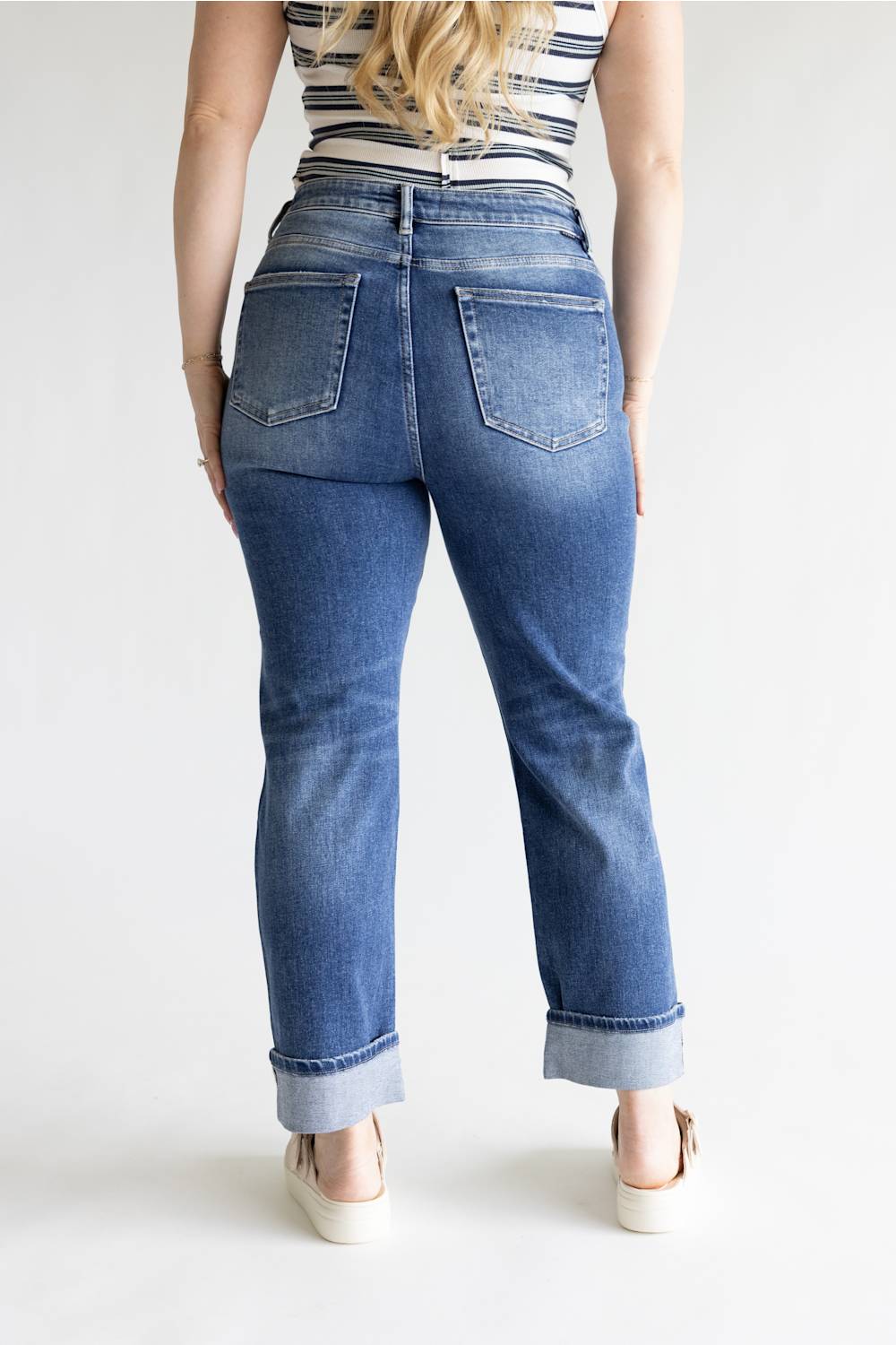 A woman viewed from the back wearing blue denim jeans with a straight-to-medium leg and rolled cuffs, paired with a striped top and white platform sandals, against a plain white studio background.
