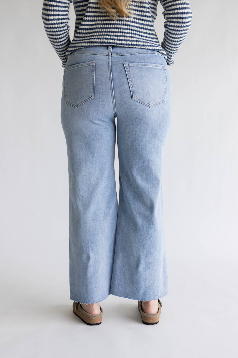 A person shown from the back wearing light-wash wide-leg denim jeans, a navy and white striped long-sleeve top, and tan flat shoes against a plain light gray studio background.
