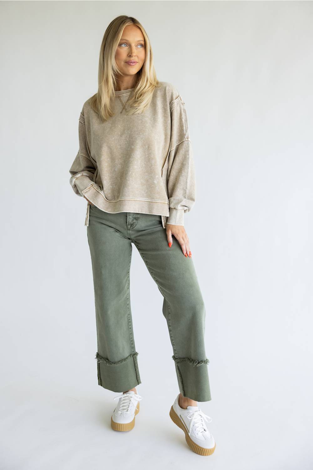 A woman modeling a beige crewneck sweatshirt, olive green cropped jeans with rolled cuffs, and white platform sneakers against a plain white studio background.