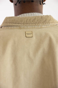 Close-up back view of a person wearing a beige overshirt or jacket with a ribbed collar and a small rectangular brand patch reading 'INDUSTRIE SUPPLY' centered below the collar.