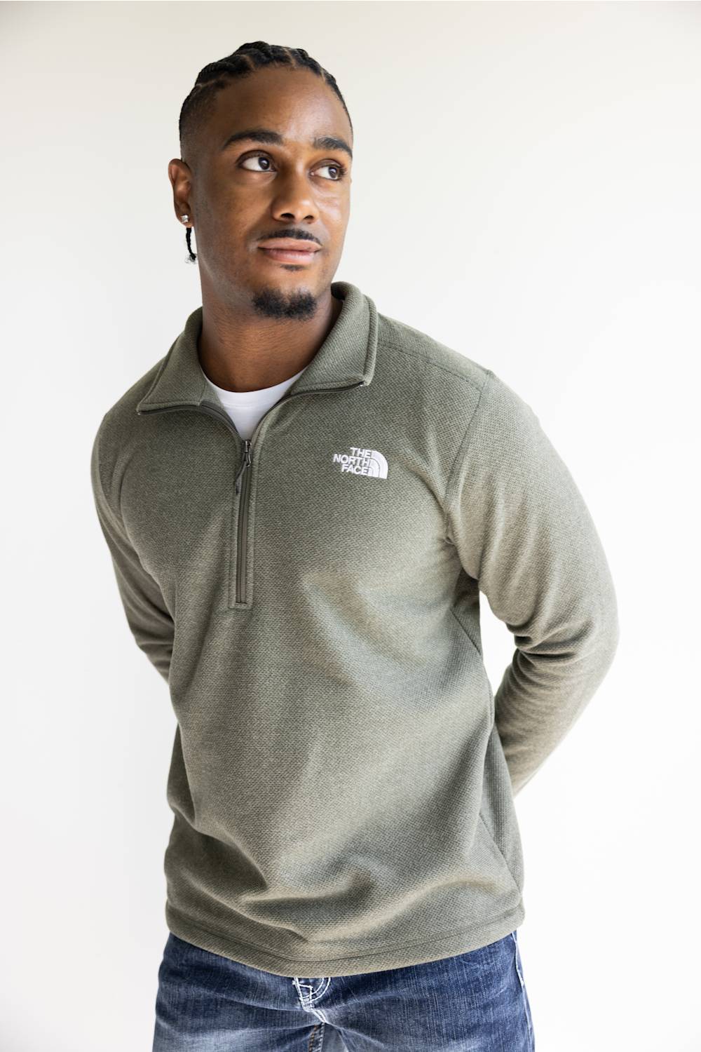 A man wearing an olive green quarter-zip pullover sweater by The North Face, paired with a white t-shirt and blue jeans, photographed against a plain light background.