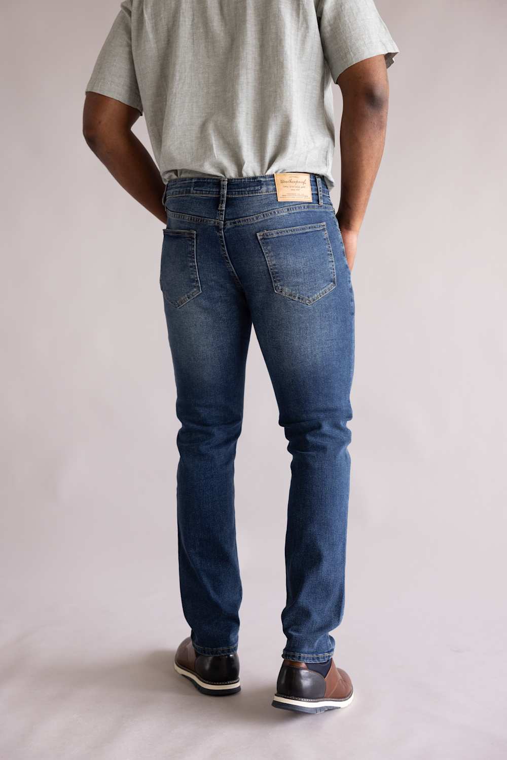 Back view of a person wearing blue jeans and a gray shirt on a plain background. The jeans are Weatherproof Vintage Slim Jeans for Men in Tidepool Tint.