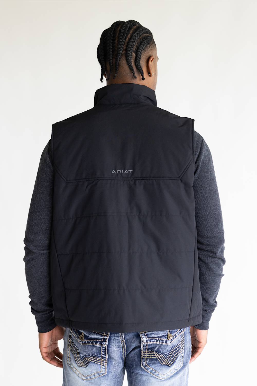 A man viewed from behind wearing a black puffer vest over a dark gray long-sleeve shirt and blue jeans. The brand name 'ARIAT' is printed in small white letters on the upper back of the vest.