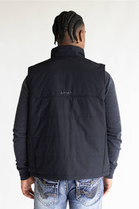 A man viewed from behind wearing a black puffer vest over a dark gray long-sleeve shirt and blue jeans. The brand name 'ARIAT' is printed in small white letters on the upper back of the vest.