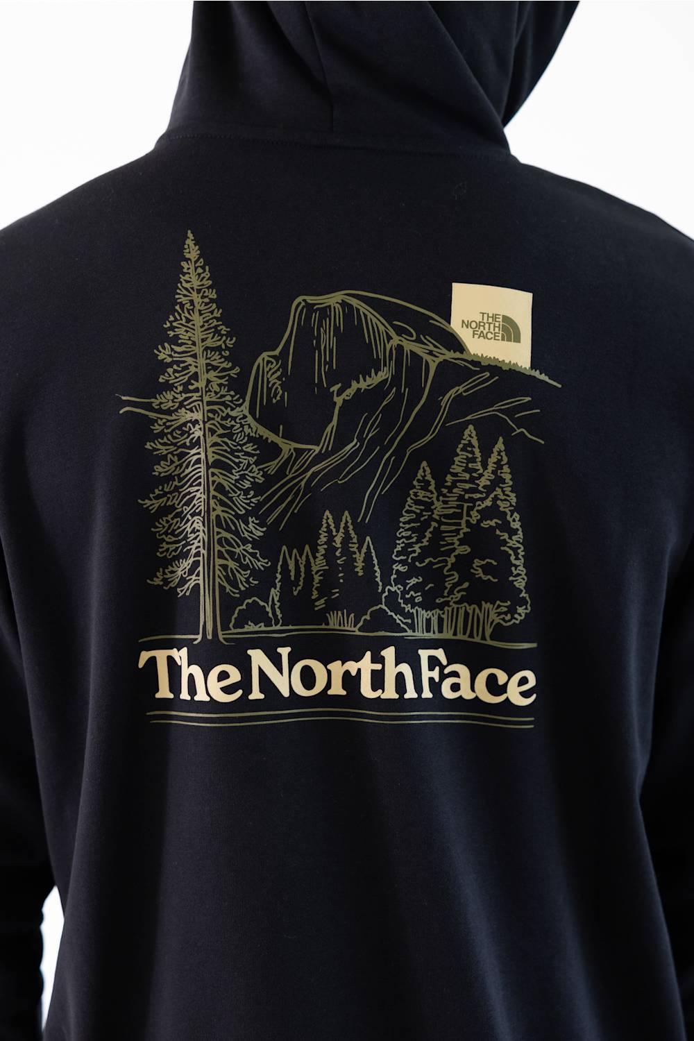 Close-up of the back of a black hoodie featuring a large gold line art graphic of a mountain and pine trees, with 'The North Face' text printed below and a small brand patch on the upper back.