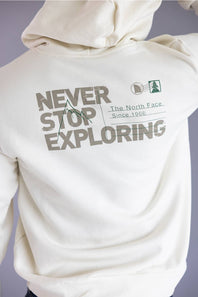 Close-up of the back of an off-white hoodie featuring a large graphic print that reads 'NEVER STOP EXPLORING' with 'The North Face Since 1966' and a small mountain-and-tree logo, worn by a person against a soft lavender-gray background.
