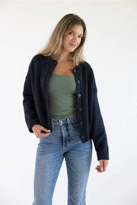 Front view of a female model wearing the open-front Things Between Alyssa Cardigan in Navy, worn unbuttoned over a sage green cami top and high-waisted blue jeans.