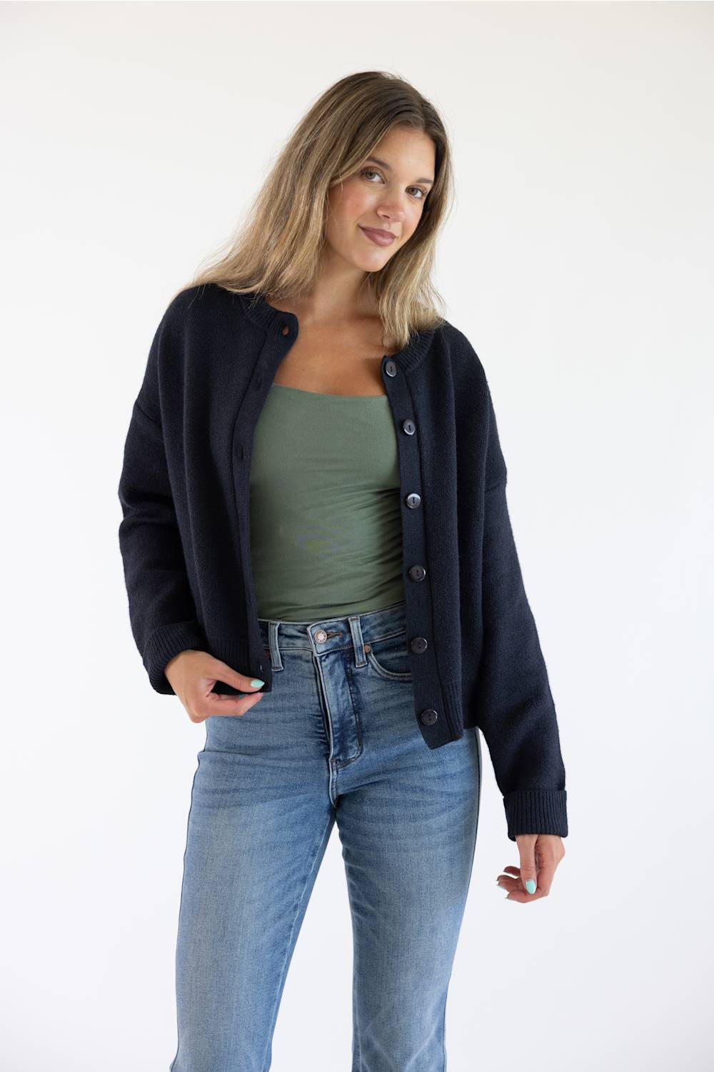 Front view of a female model wearing the open-front Things Between Alyssa Cardigan in Navy, worn unbuttoned over a sage green cami top and high-waisted blue jeans.