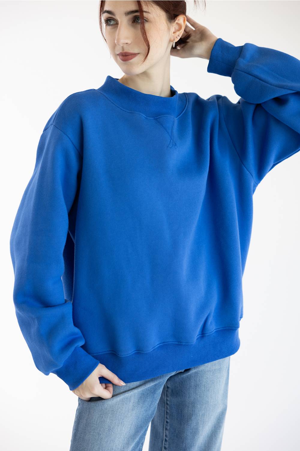 A woman wearing a vibrant royal blue crewneck sweatshirt and light wash blue jeans, posing against a clean white studio background.