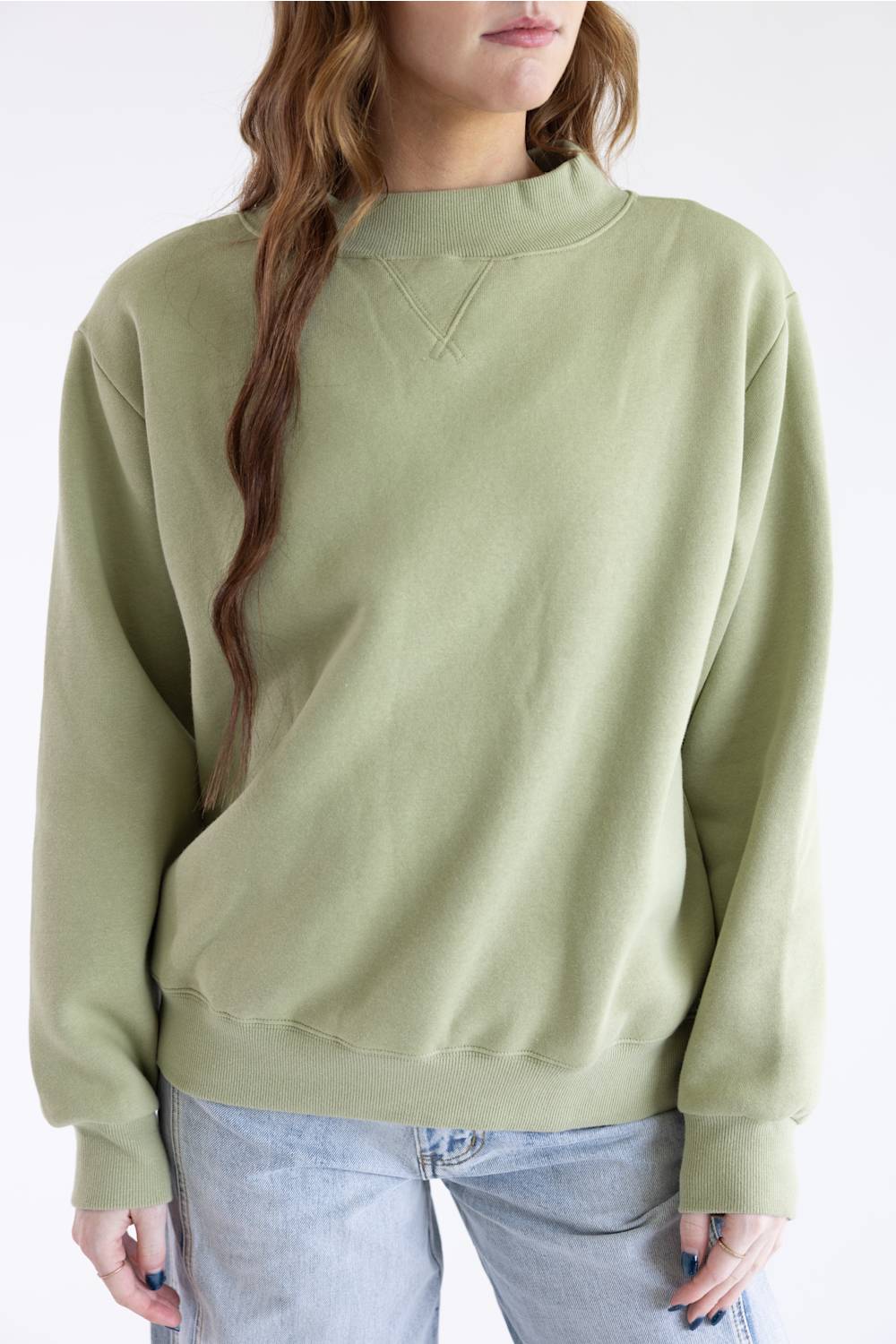 A woman wearing an oversized sage green crewneck sweatshirt and light blue jeans, shown from the chest down against a plain white background.