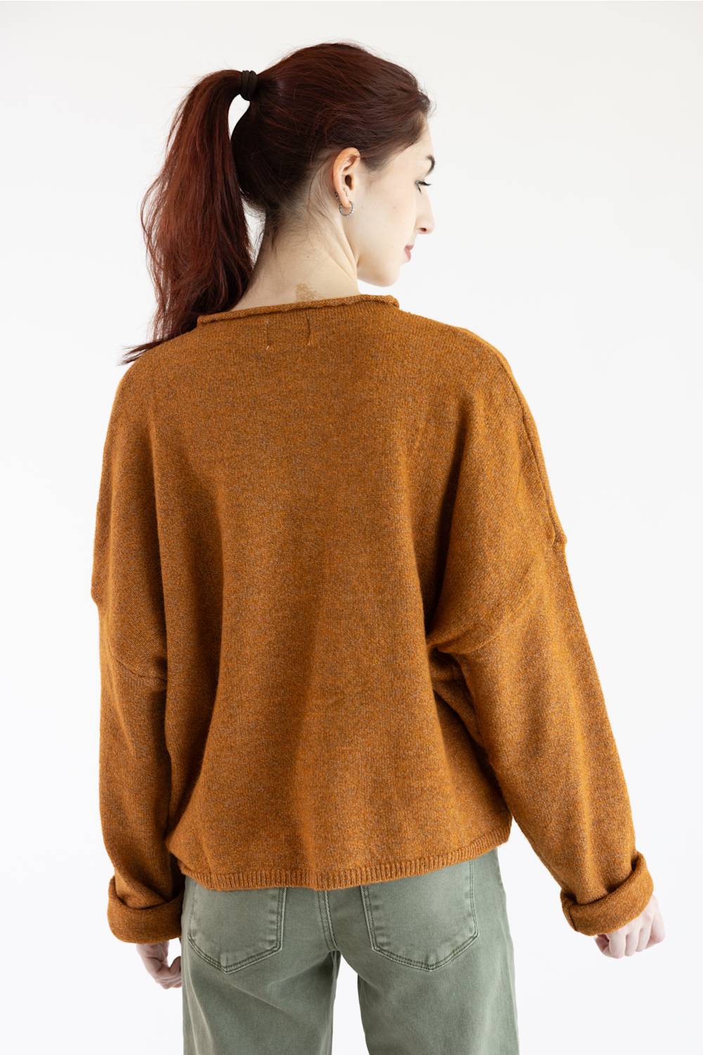 A woman with auburn hair in a ponytail is shown from the back wearing a loose-fit, textured knit sweater in a warm rust-brown color, paired with olive green pants, against a plain white studio background.
