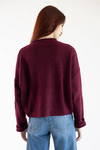 A woman viewed from behind wearing a dark burgundy knit cardigan sweater and blue jeans against a plain white background.