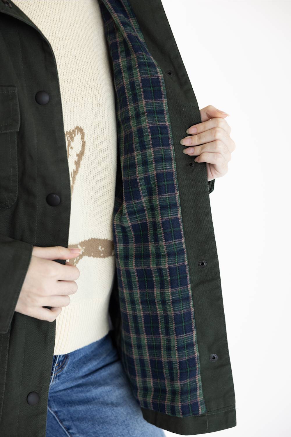 A close-up of a person wearing an open dark olive green jacket over a cream knit sweater and blue jeans, showing the interior lining of the jacket in a green and navy plaid pattern.