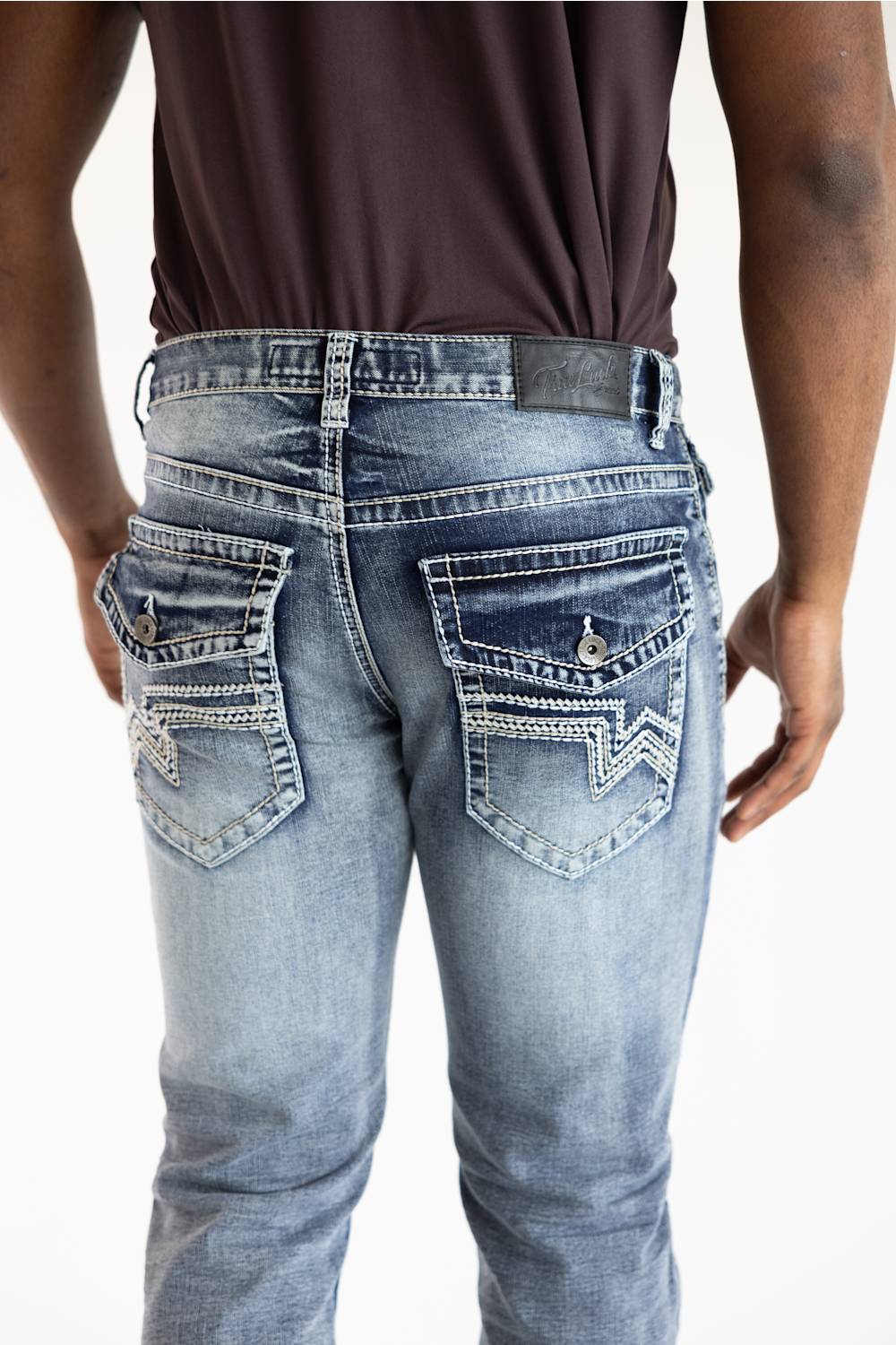 Rear view of a person wearing light-wash blue denim jeans with decorative contrast stitching and zigzag embroidery on the back pockets, paired with a dark brown shirt against a white background.