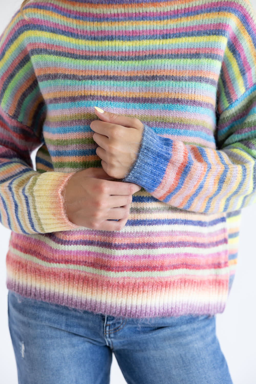 Close up of a person wearing a striped multi rainbow colored long sleeve sweater with blue denim jeans standing in front of a white background