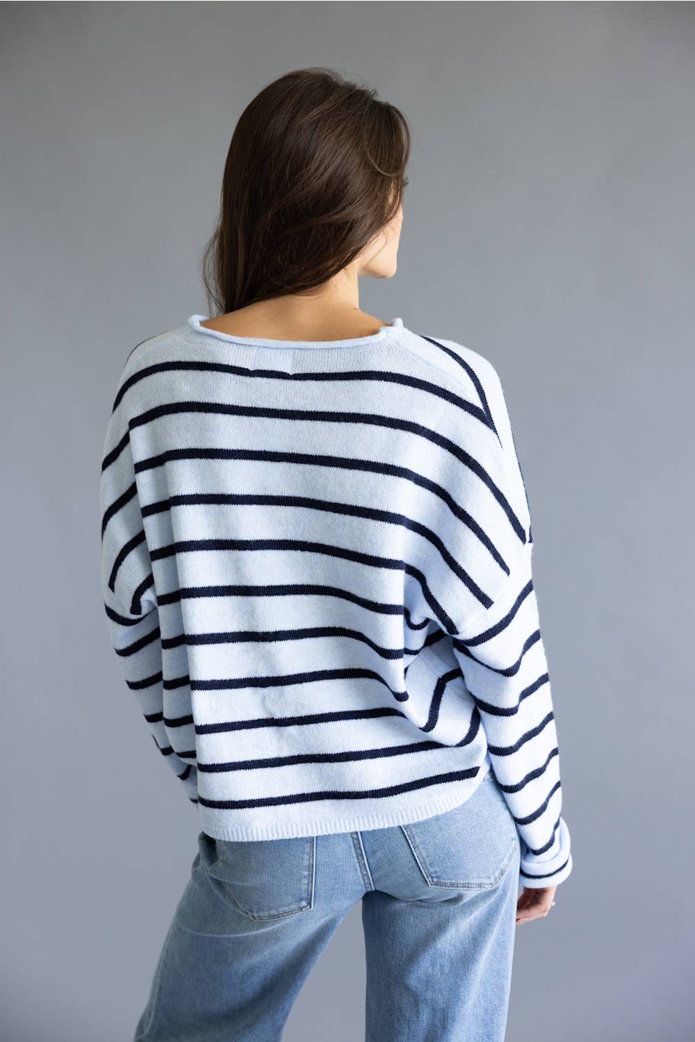 A woman viewed from behind wearing a white knit sweater with horizontal navy stripes and light blue jeans, set against a plain gray studio background.