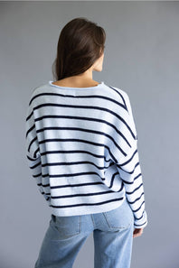 A woman viewed from behind wearing a white knit sweater with horizontal navy stripes and light blue jeans, set against a plain gray studio background.