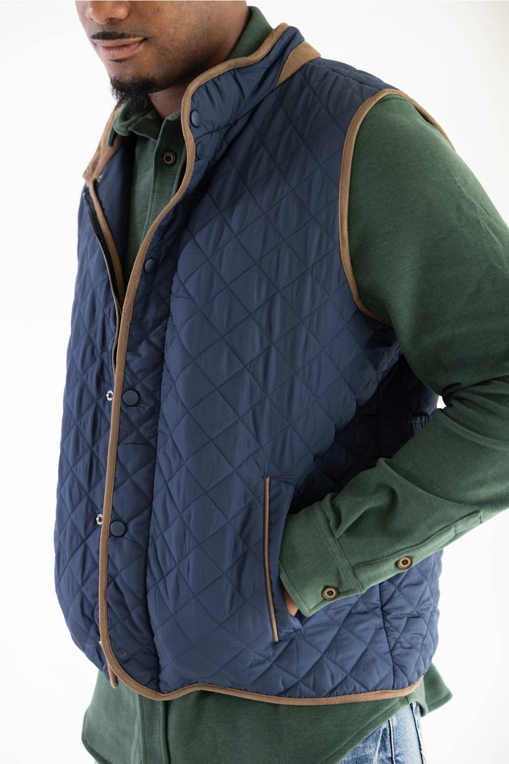 A man wearing a navy blue diamond-quilted vest with tan trim over a green long-sleeve shirt, shown from the chest down against a plain white background.