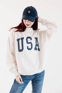 A woman wearing a cream-colored turtleneck sweatshirt with 'USA' in navy blue polka-dot letters on the front, paired with blue jeans and a navy blue baseball cap with a white NY logo.