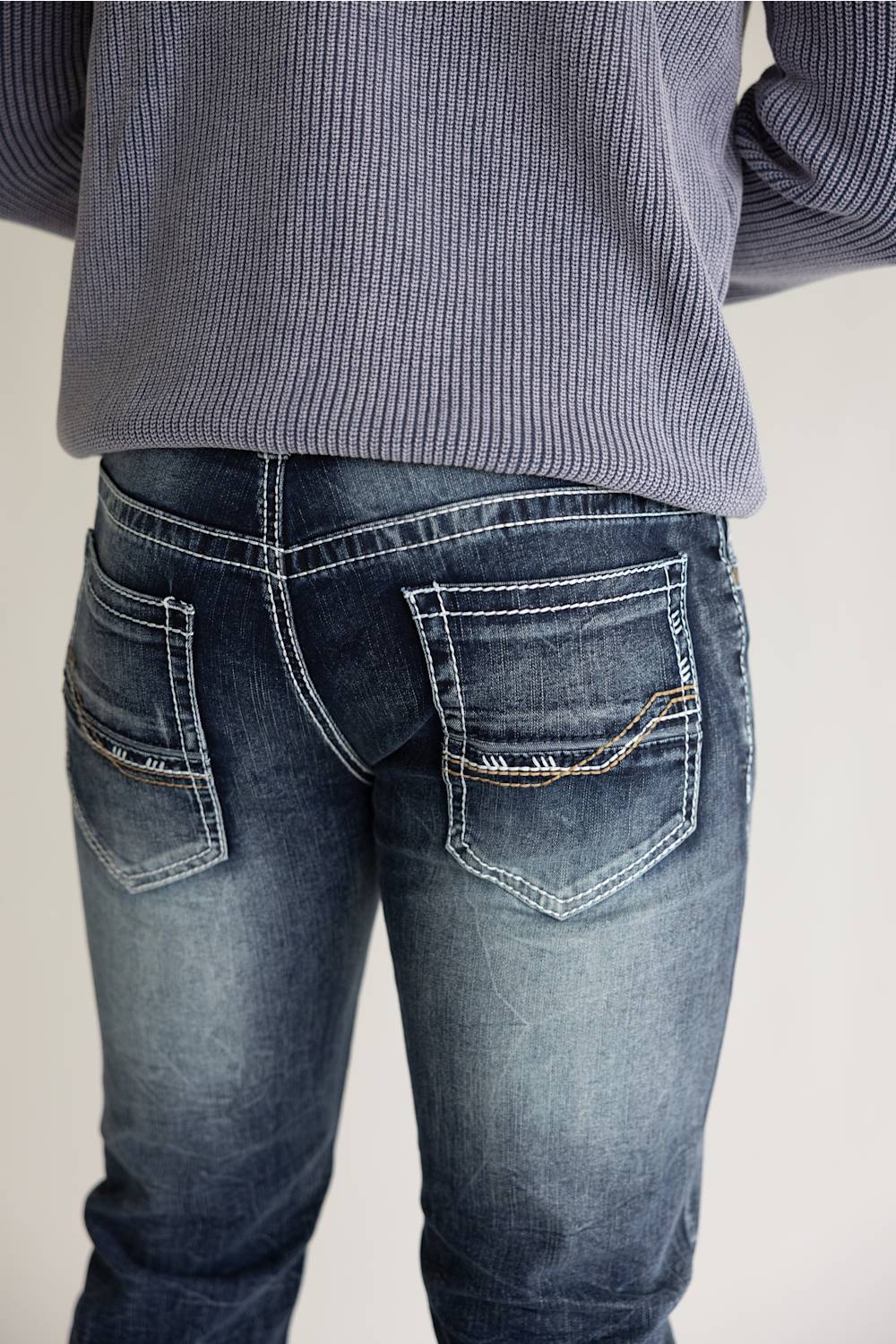 Rear view of a person wearing dark blue denim jeans with contrast stitching and a ribbed gray top, shown against a plain light background.
