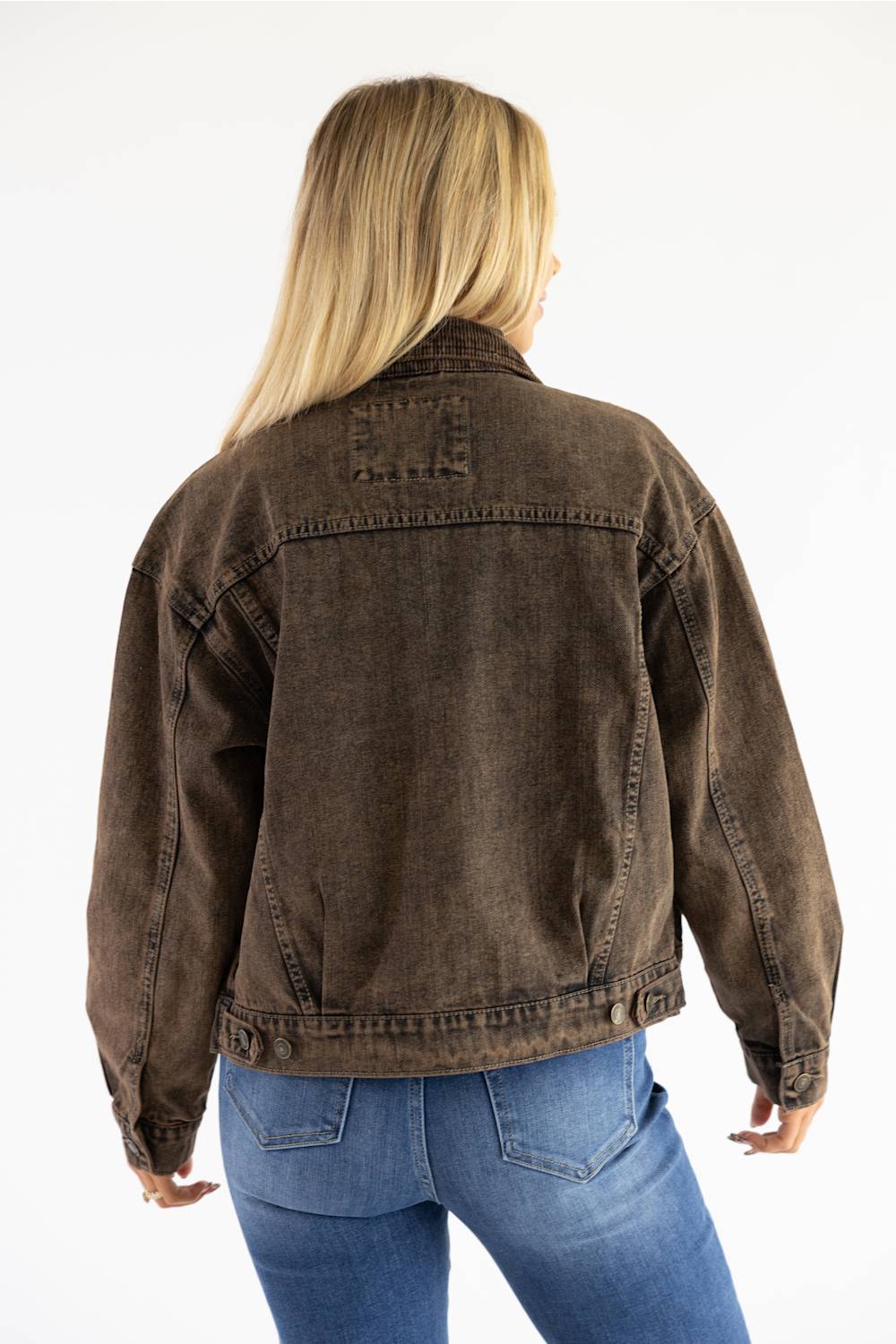 A woman viewed from the back wearing a dark brown washed denim jacket and blue jeans against a plain white background.