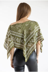 Back view of a model wearing an olive green asymmetric scarf poncho top featuring a white floral, scroll, and polka dot border print, paired with black leather pants against a plain white studio background.