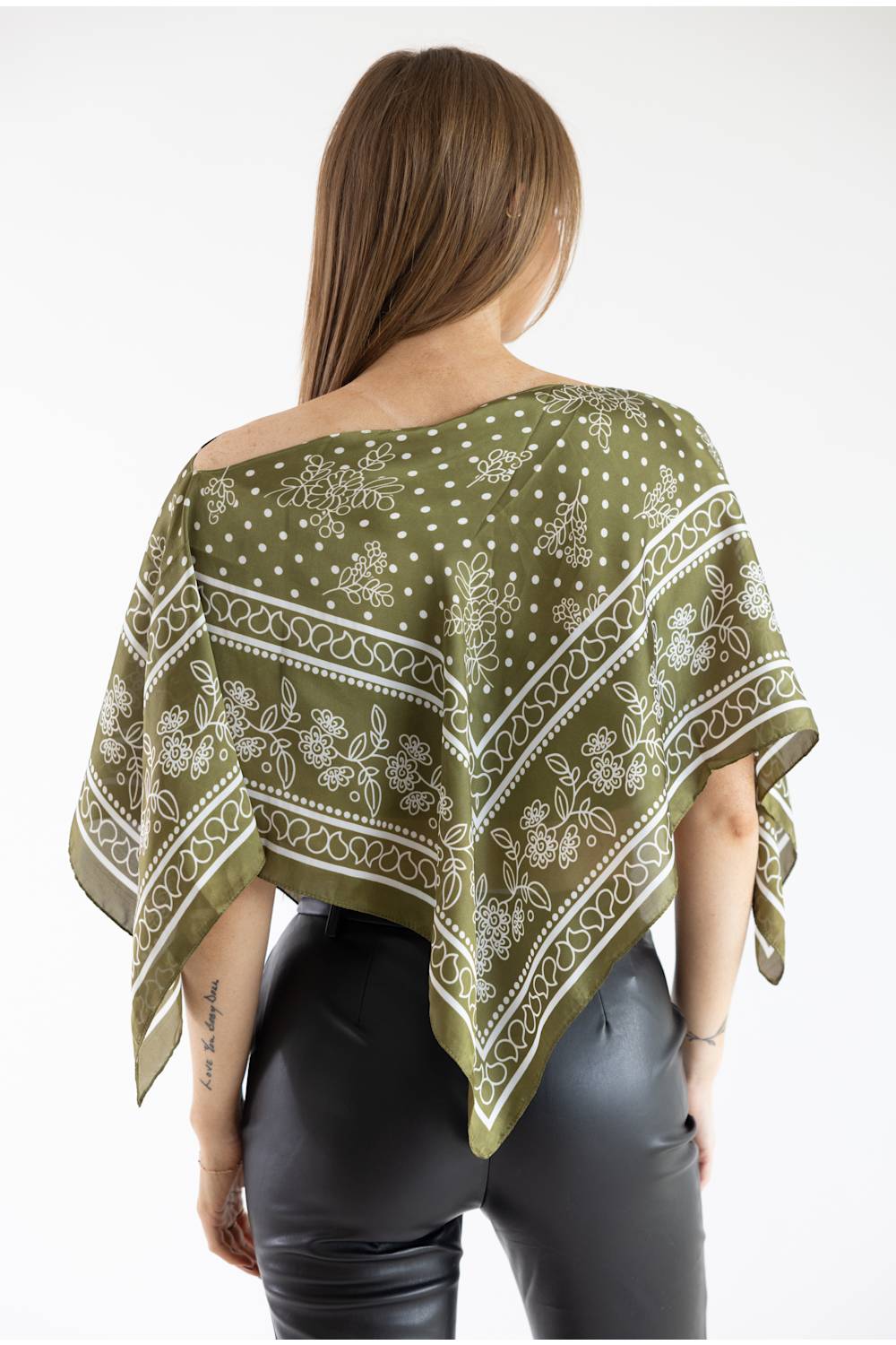 Back view of a model wearing an olive green asymmetric scarf poncho top featuring a white floral, scroll, and polka dot border print, paired with black leather pants against a plain white studio background.