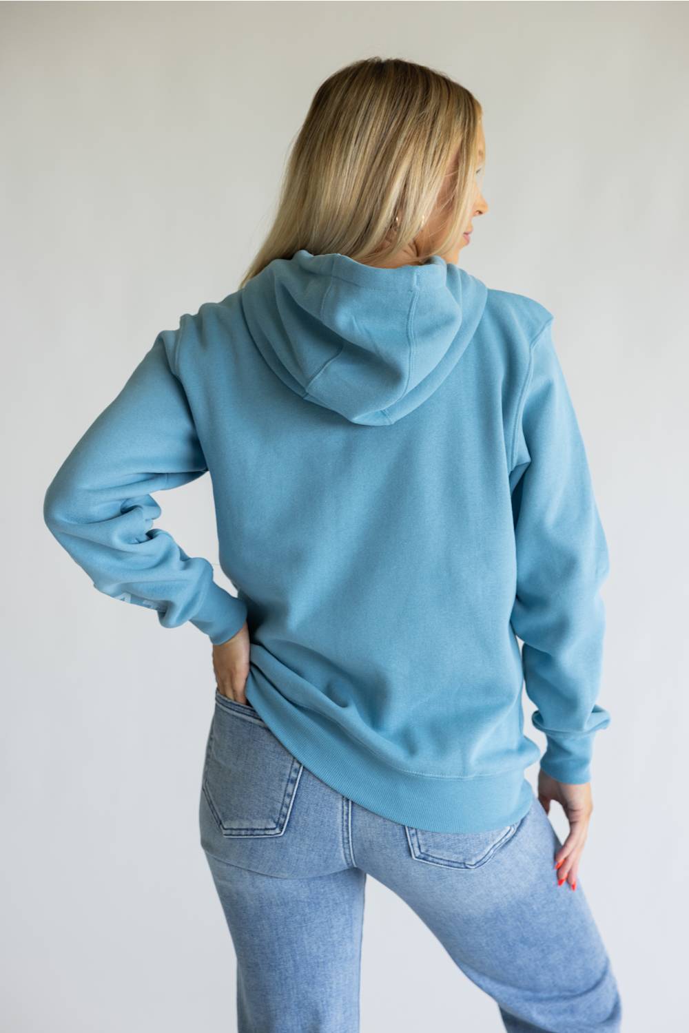 A woman with blonde hair is shown from behind wearing a dusty blue pullover hoodie and light wash blue jeans against a plain white background.