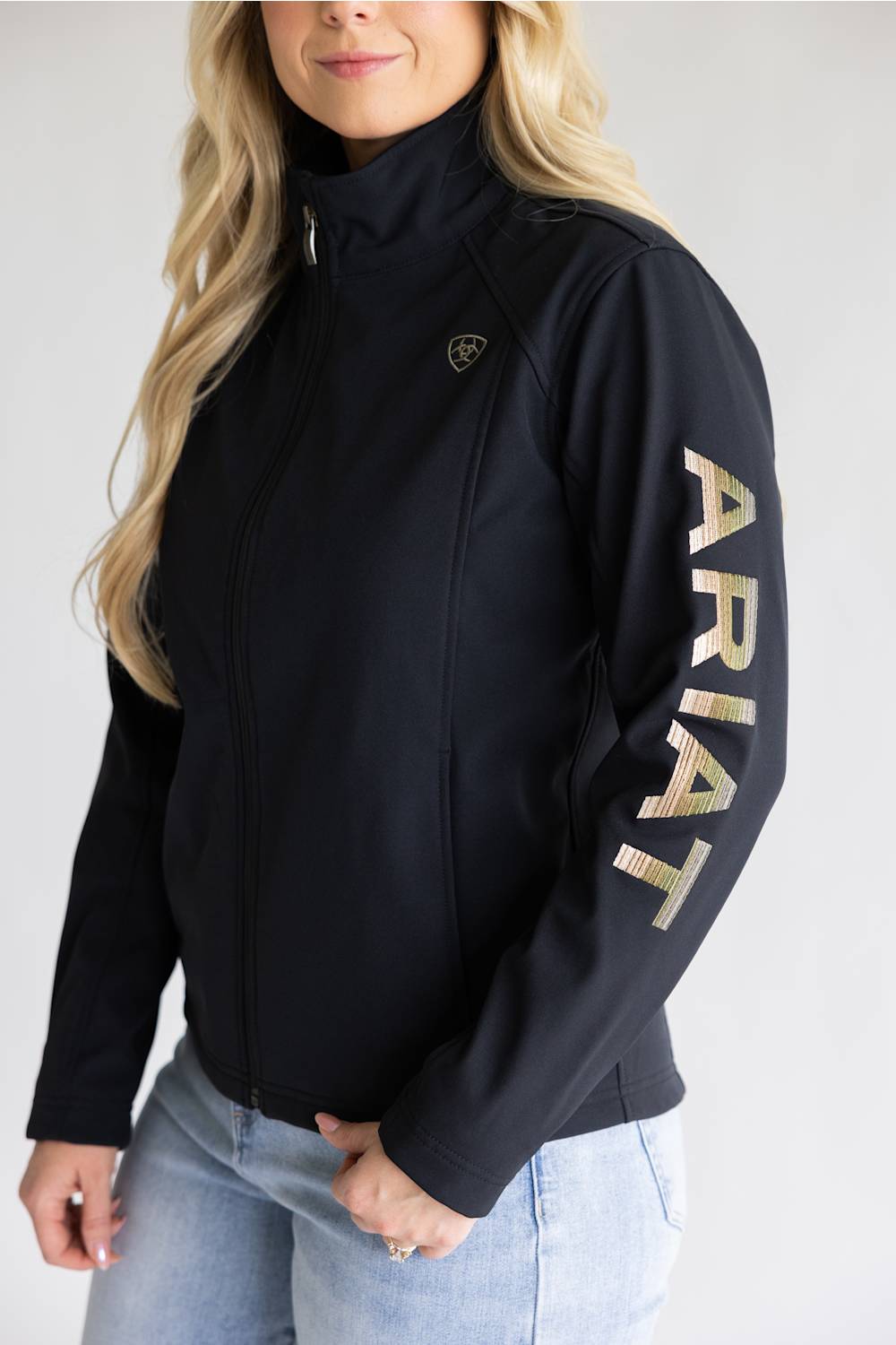 A woman wearing a black zip-up jacket with a high collar and large metallic gold 'ARIAT' lettering down one sleeve, paired with light blue jeans against a light gray studio background.