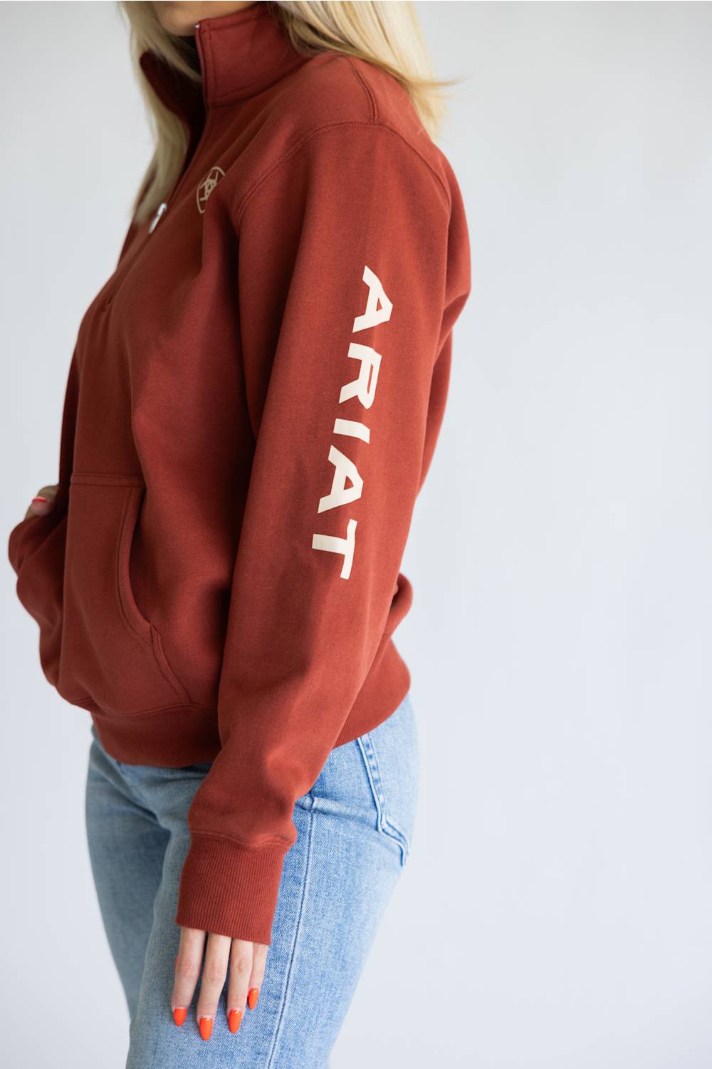 A woman wearing a rust-colored quarter-zip hoodie with 'ARIAT' printed in white down the left sleeve, paired with light blue jeans against a plain light gray background.