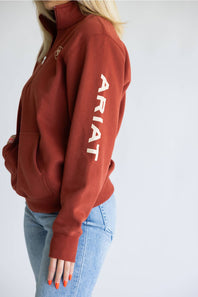 A woman wearing a rust-colored quarter-zip hoodie with 'ARIAT' printed in white down the left sleeve, paired with light blue jeans against a plain light gray background.