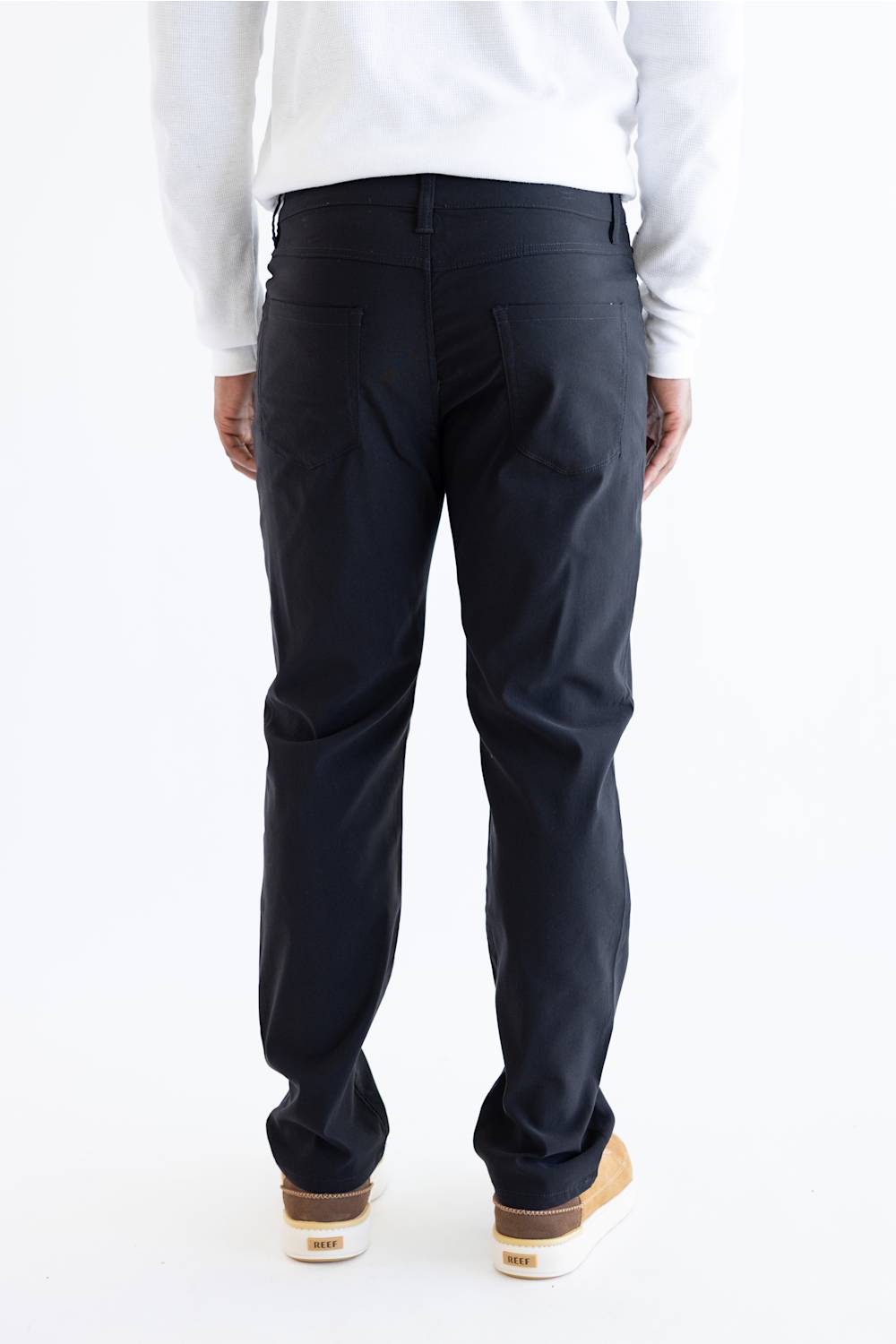 Rear view of a person wearing dark navy pants, a white long-sleeve top, and tan-and-white sneakers against a plain white studio background.