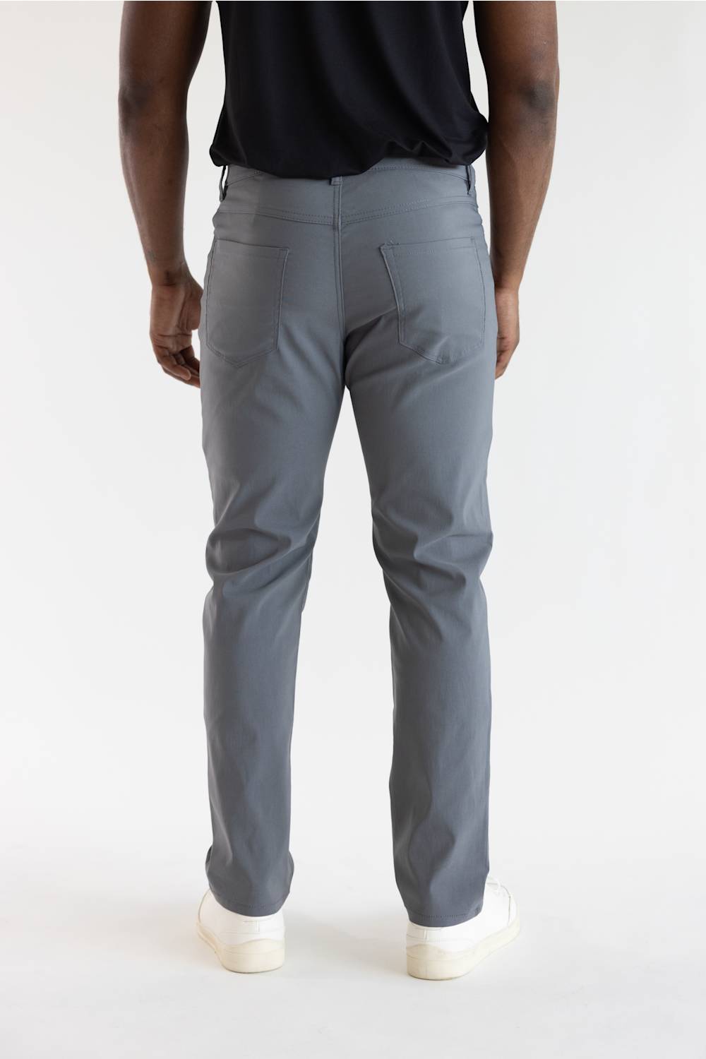 A person shown from the back wearing gray slim-fit trousers, a black top, and white sneakers against a plain light background.