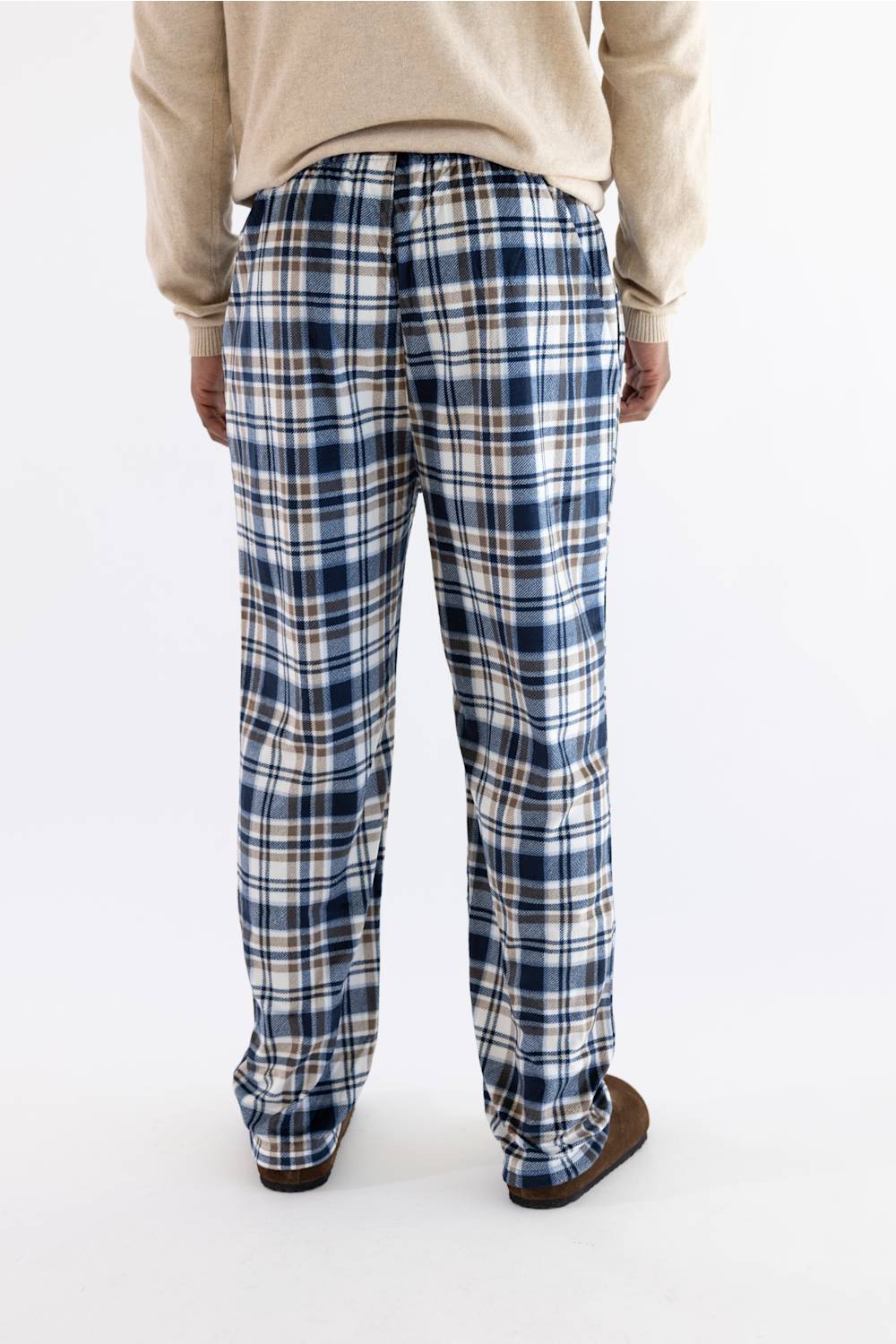 A person shown from the back wearing loose plaid pants with a navy, white, and beige/tan pattern, paired with a beige sweater and brown slippers against a plain white studio background.