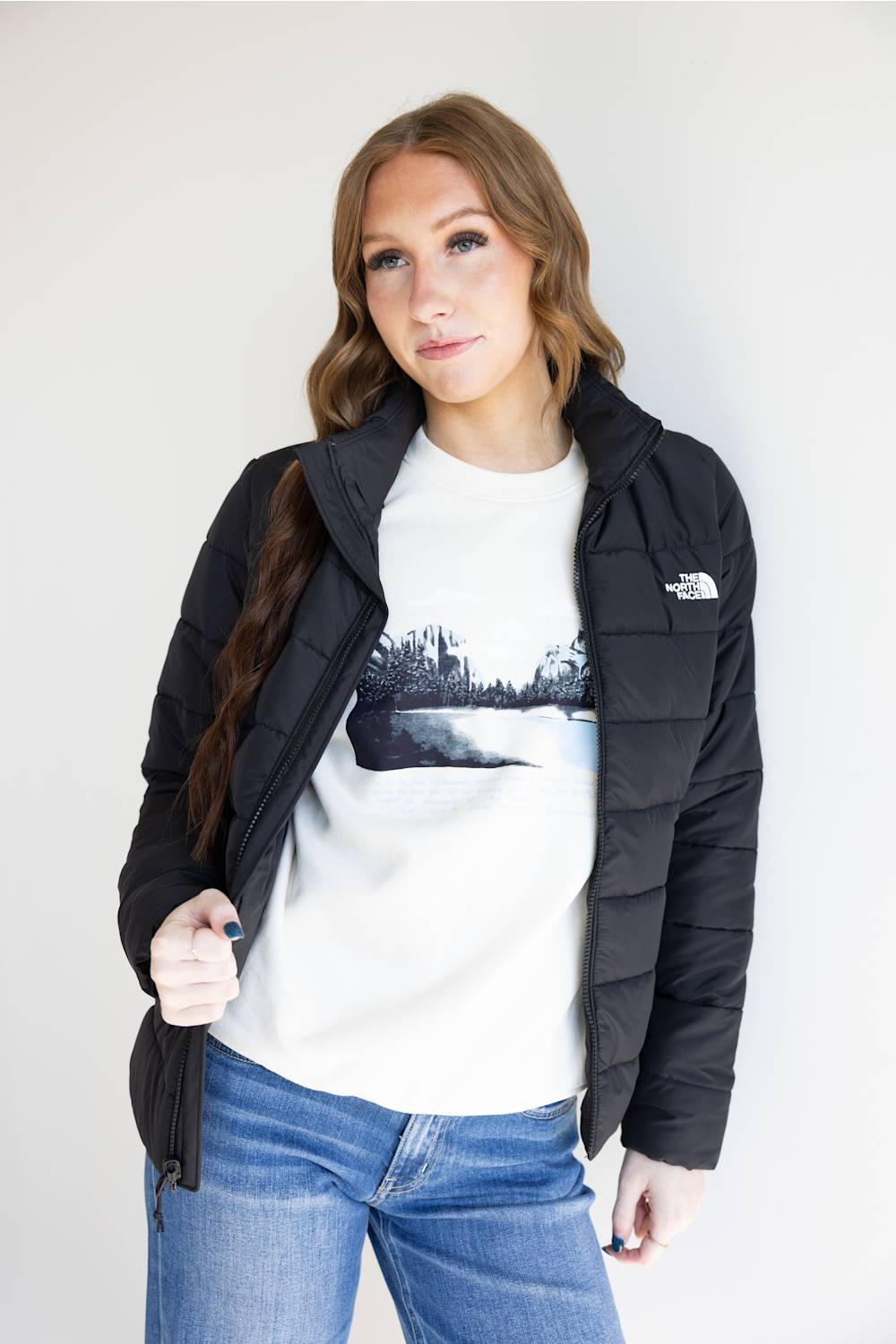 A woman wearing a black puffer jacket by The North Face over a white graphic t-shirt and blue jeans, standing against a plain light gray background.