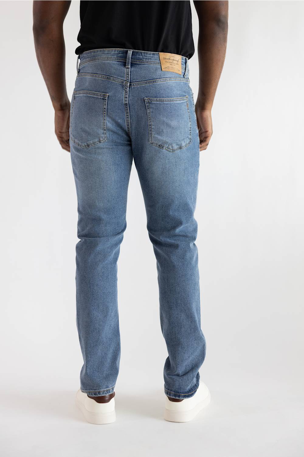 Rear view of a person wearing light blue denim jeans, a black t-shirt, and white sneakers against a plain white studio background.