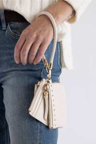 A close-up of a person wearing blue jeans and a cream knit sweater, holding a small cream leather shoulder bag with gold-tone hardware and a matching wrist strap.