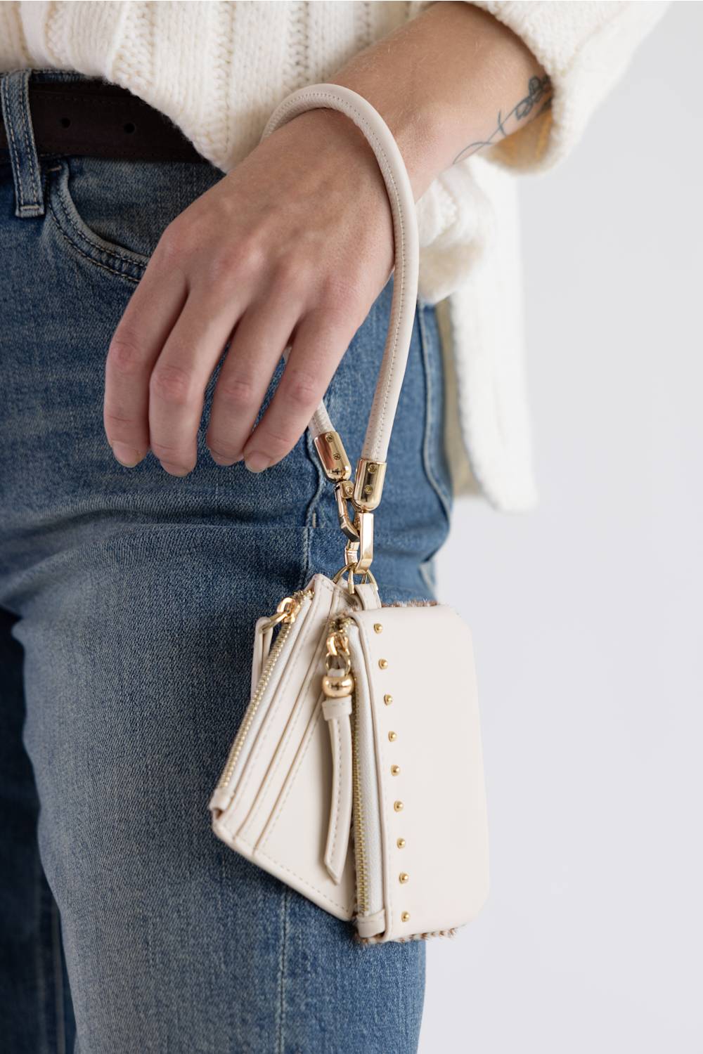 A close-up of a person wearing blue jeans and a cream knit sweater, holding a small cream leather shoulder bag with gold-tone hardware and a matching wrist strap.