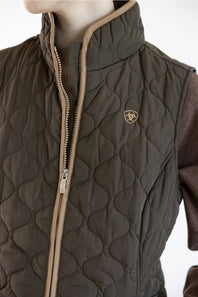 A close-up of a quilted brown vest with a stand-up collar, tan zipper trim, and a small shield logo on the chest, worn over a brown long-sleeve shirt against a white background.