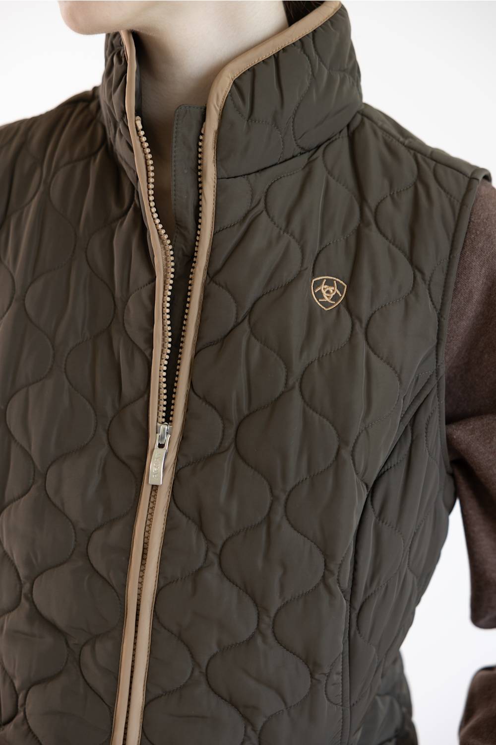 A close-up of a quilted brown vest with a stand-up collar, tan zipper trim, and a small shield logo on the chest, worn over a brown long-sleeve shirt against a white background.