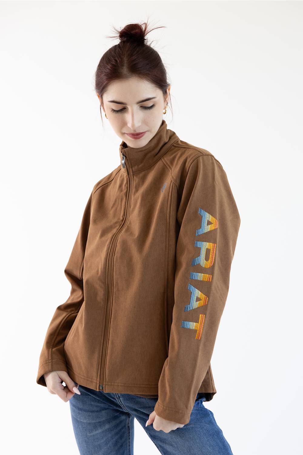 A woman wearing a brown zip-up jacket with a colorful ARIAT logo printed vertically on the sleeve, paired with blue jeans, against a plain white background.