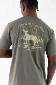 A man viewed from behind wearing a sage green Carhartt t-shirt with a large beige graphic on the back featuring a deer, trees, and the Carhartt logo.