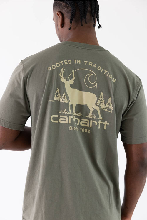 A man viewed from behind wearing a sage green Carhartt t-shirt with a large beige graphic on the back featuring a deer, trees, and the Carhartt logo.